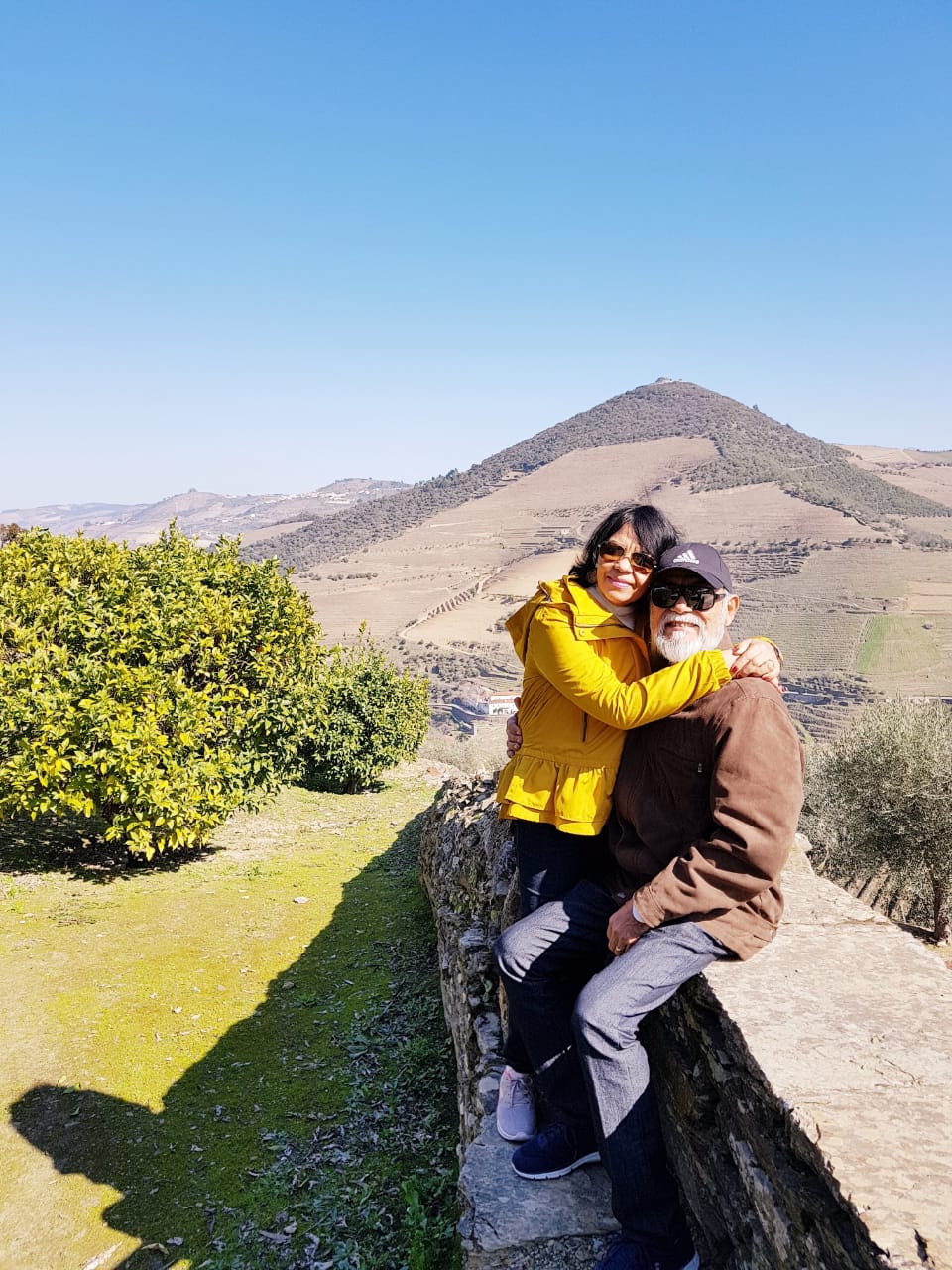  Foto de Carlos e sua esposa sentados em um muro em uma paisagem montanhosa