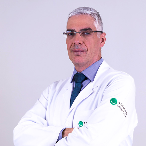 Doutor Sacomani, branco, cabelo branco, jaleco branco, óculos preto, sorri de braços cruzados