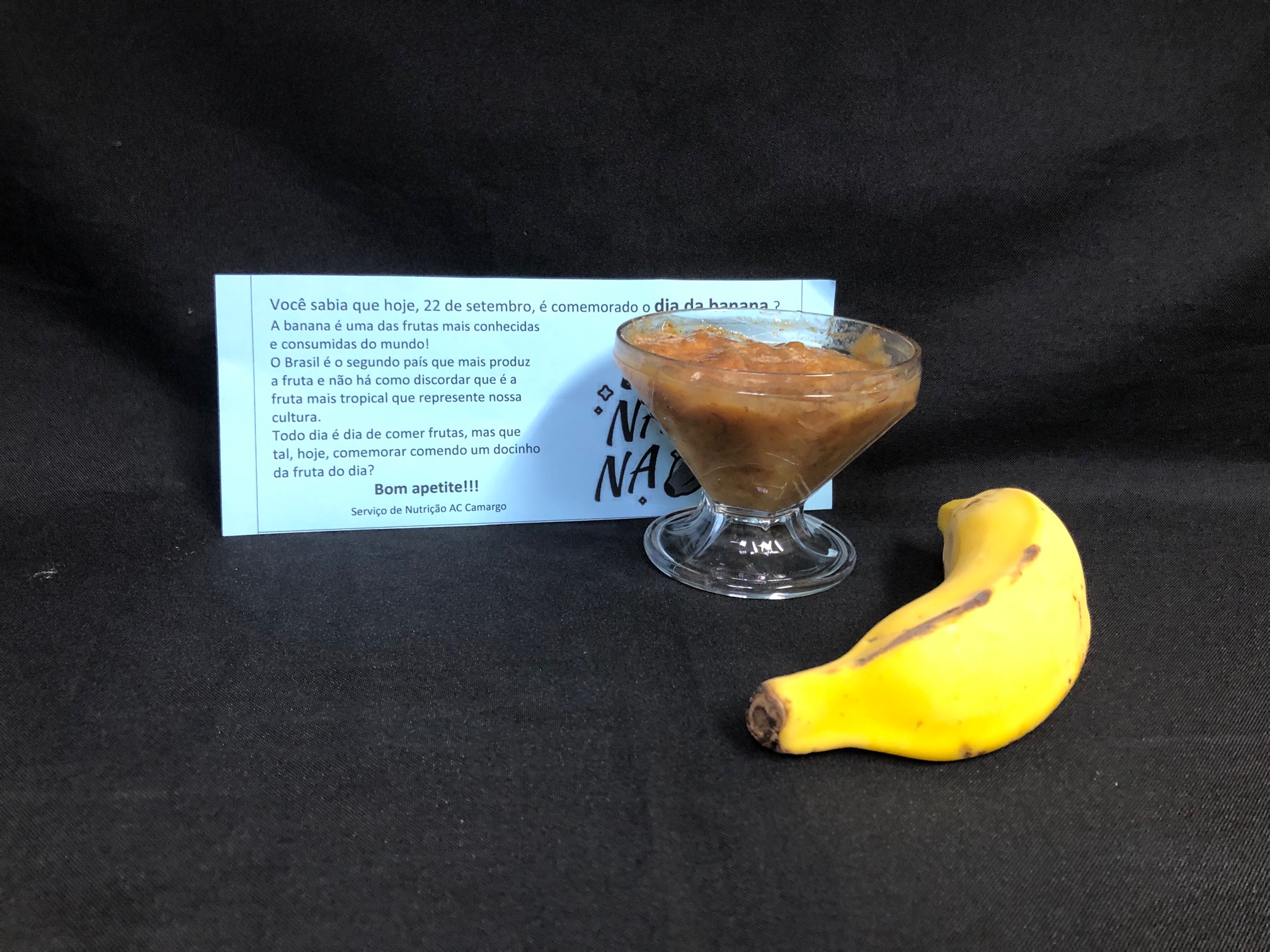 taça de vidro transparente com o doce de banana, pastoso e marrom; ao lado, uma banana, comprida e amarela