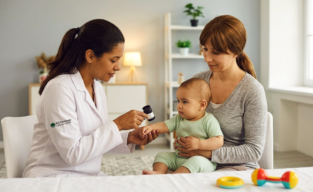 Médica examinando com uma lupa dermatológica o antebraço de um bebê que está deitado no colo da mãe, em consultório claro.
