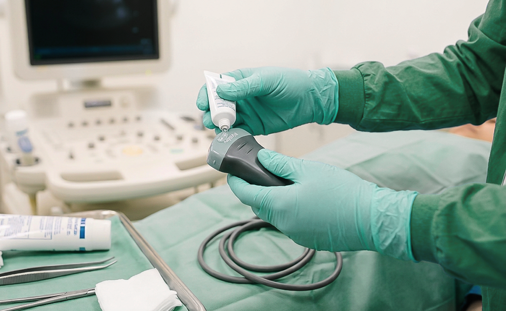 Mãos enluvadas ajustando um transdutor de ultrassom sobre campo cirúrgico estéril, com bandeja de instrumentos ao lado.
