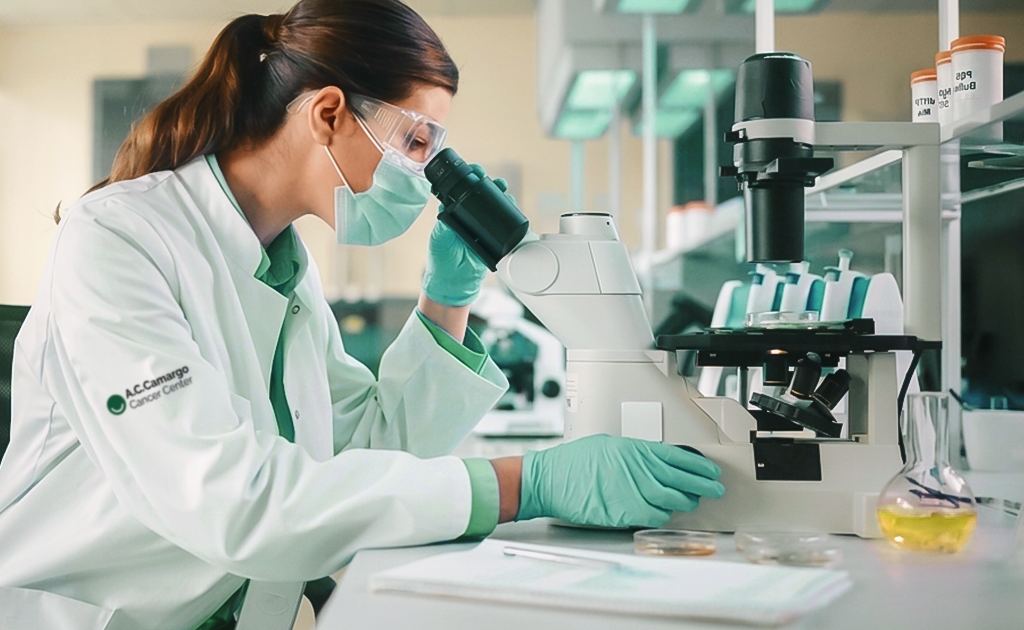 Patologista ajustando o foco de um microscópio duplo em laboratório claro, com uma bandeja de lâminas de vidro sobre a bancada.