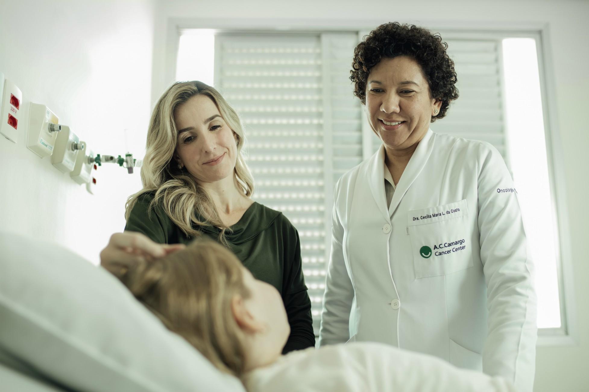 Dra. Cecilia Maria Lima da Costa olha sorrindo para a garota na cama do hospital, ao lado da mãe dela, em pé