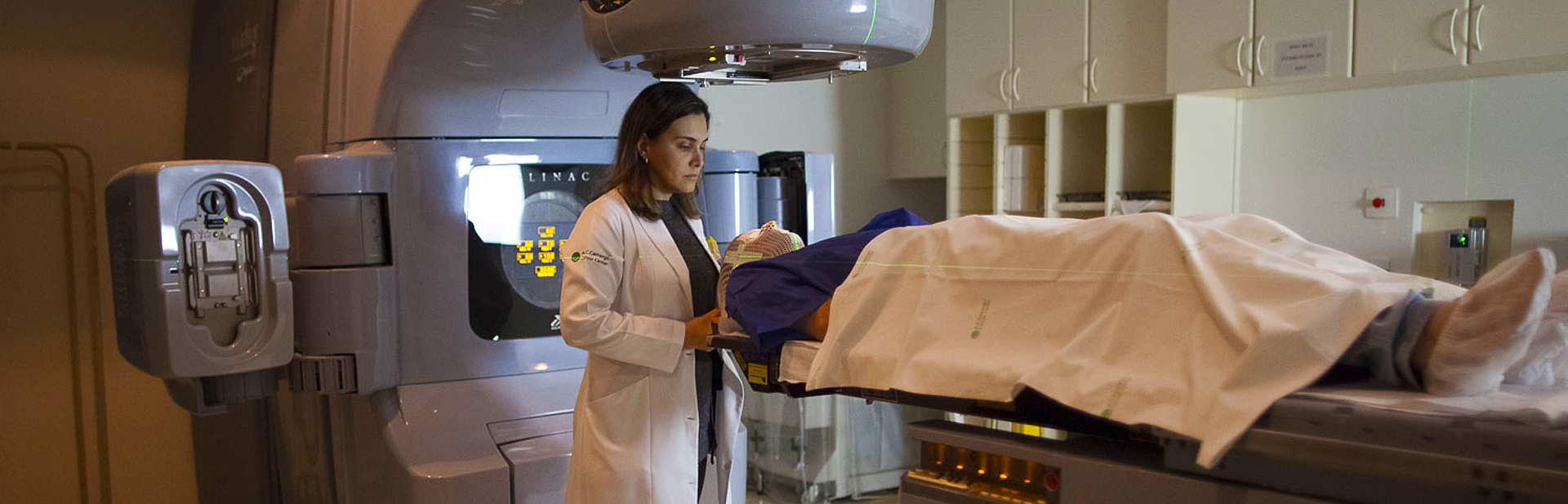 Uma médica branca de cabelos castanhos lisos ajustando um equipamento de radioterapia na cabeça de um paciente