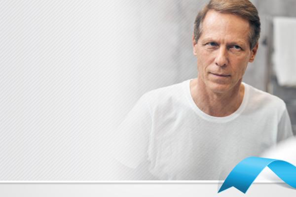 Homem branco de olhos azuis usando camiseta branca se olhando no espelho, no canto inferior direito, um laço azul colocado digitalmente na imagem