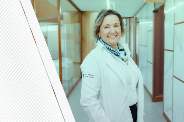 Doutora Solange, cabelos curtos e loiros, sorri em pé com o jaleco do hospital