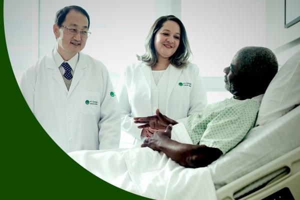 Paciente idoso e negro deitado na cama de hospital; em pé, um médico japonês e uma enfermeira morena sorriem
