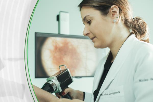 Doutora Juliana Casagrande, branca e com cabelos castanhos lisos, fazendo o exame de dermatoscopia em montagem com o laço laranja da campanha Dezembro Laranja