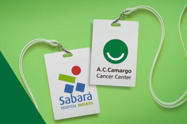 Dois crachás brancos com o logo do Hospital Sabará Infantil e do A.C.Camargo Cancer Center. O fundo é uma montagem de duas formas geométricas em dois tons de verde.