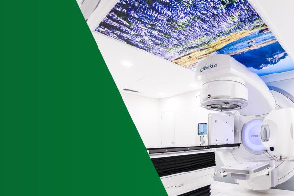 O Acelerador Linear, uma máquina branca com uma maca para fazer radioterapia; no teto, uma paisagem de campo para relaxar pacientes