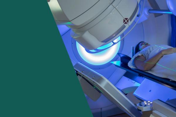 Mulher jovem branca deitada numa máquina branca de radioterapia, há objeto redondo com luz azul iluminando sua cabeça