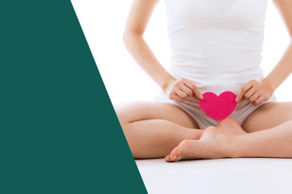 Corpo de uma mulher branca e magra do pescoço pra baixo, roupa branca; ela segura um coração rosa em frente ao seu órgão genital