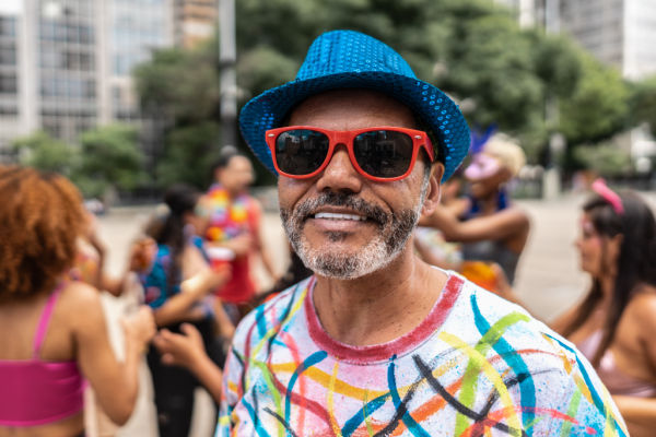 Homem no carnaval, com camisa colorida, chápeu e óculos escuros, sorrindo