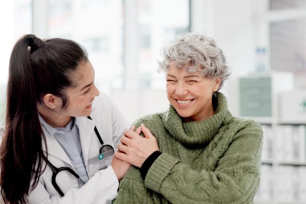 Médica mulher, sorrindo, com a mão no ombro de uma paciente mulher, também sorrindo.
