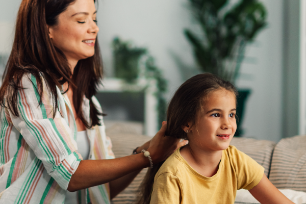 Mãe penteando o cabelo da filha e apoiando a mão na lateral do pescoço da criança, em um gesto de carinho e atenção.
