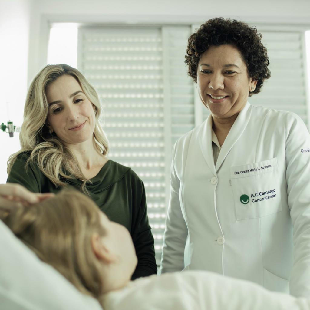 Dra. Cecilia Maria Lima da Costa olha sorrindo para a garota na cama do hospital, ao lado da mãe dela, em pé