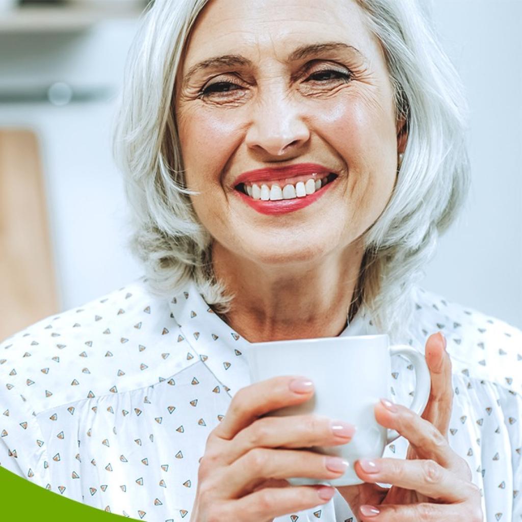 Mulher de cabelo branco sorri com seu batom vermelho e uma caneca na mão
