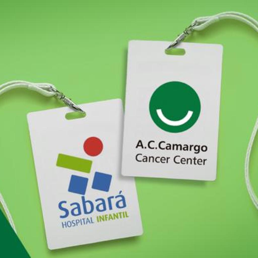 Dois crachás brancos com o logo do Hospital Sabará Infantil e do A.C.Camargo Cancer Center. O fundo é uma montagem de duas formas geométricas em dois tons de verde.