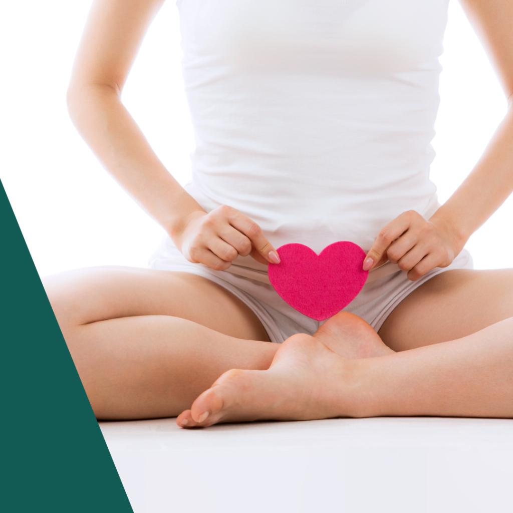 Corpo de uma mulher branca e magra do pescoço pra baixo, roupa branca; ela segura um coração rosa em frente ao seu órgão genital