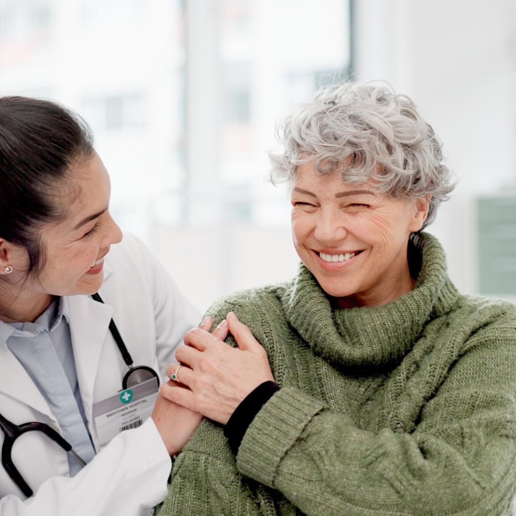 Médica mulher, sorrindo, com a mão no ombro de uma paciente mulher, também sorrindo.