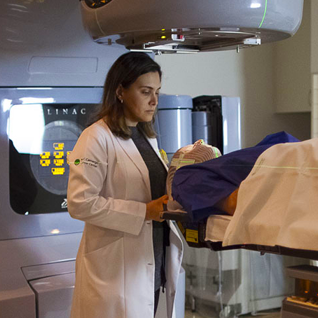 Uma médica branca de cabelos castanhos lisos ajustando um equipamento de radioterapia na cabeça de um paciente